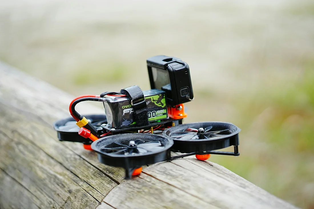 Five-inch cinewhoop FPV quadcopter with guarded propellers, battery and action camera resting outdoors.