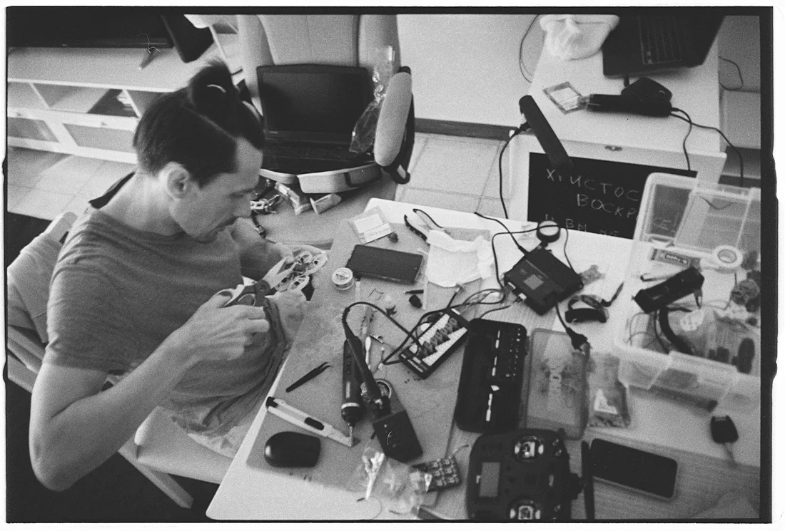 Black-and-white photograph of a drone hobbyist working on electronics at a parts-covered workbench